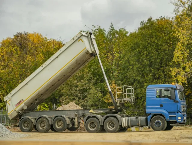 A blue dump truck unloading at a construction site surrounded by autumn foliage.