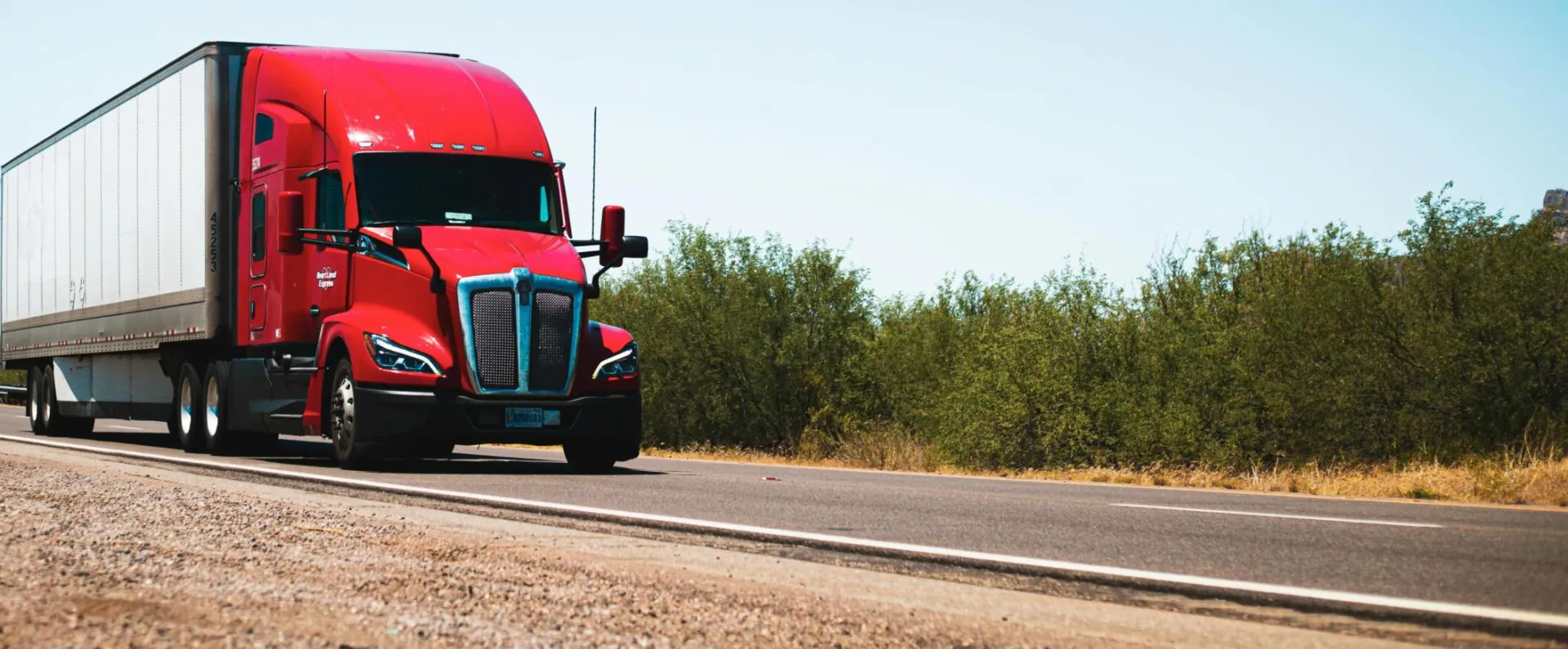 Vibrant red semi-trailer truck driving on an open highway under a clear blue sky.