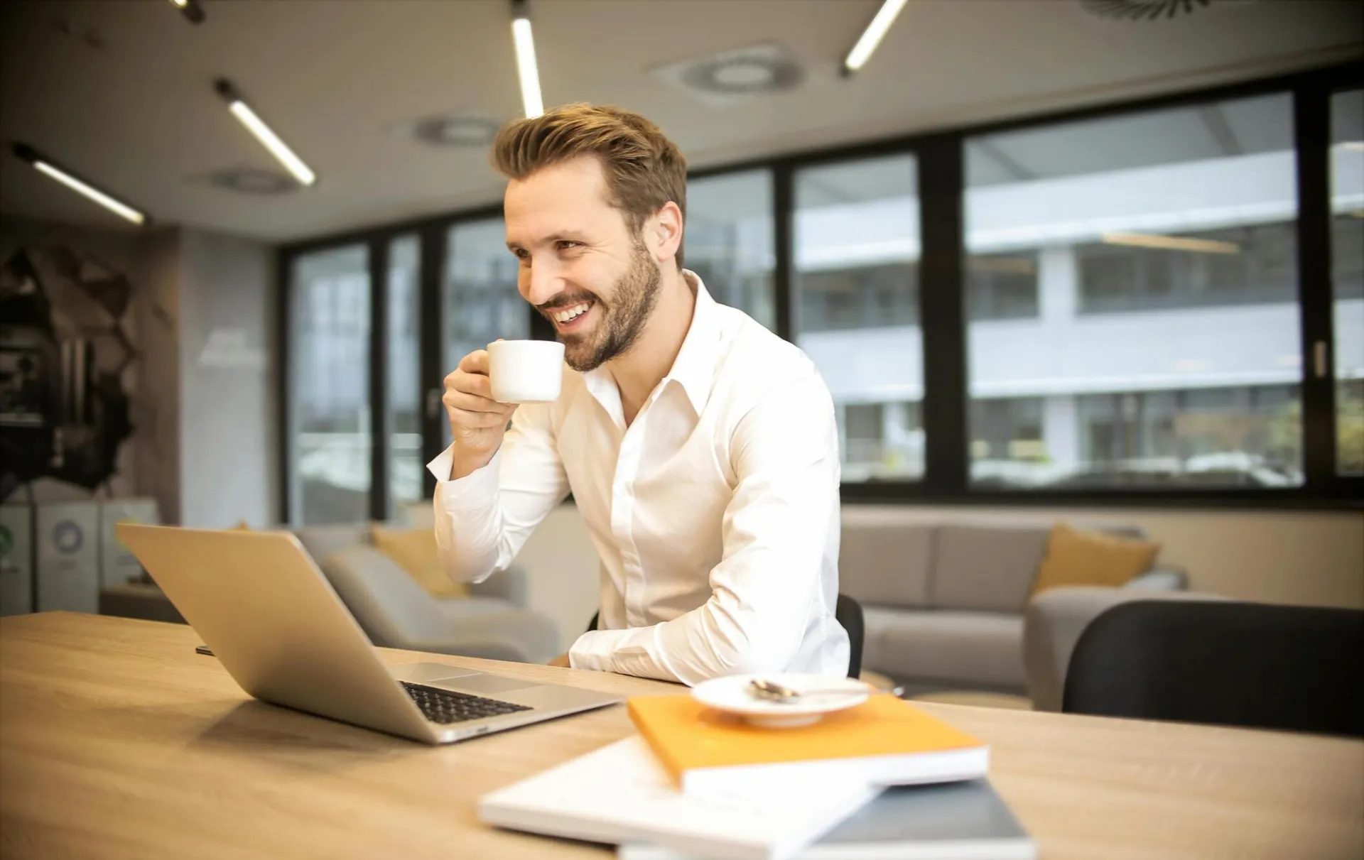 Man enjoying a coffee break while working in a modern office with laptop and books.