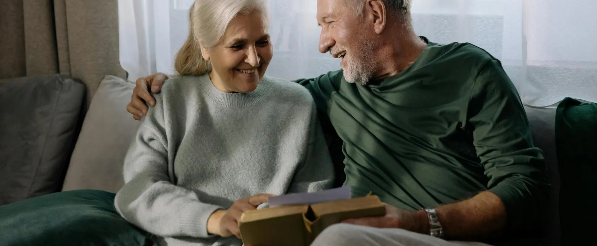 Senior couple embracing and reading a book on a sofa, sharing a joyful and intimate moment indoors.