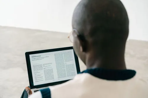Bald man reading a document on a laptop screen in a minimalist indoor setting.