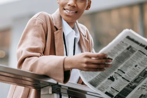 Crop happy African American businesswoman in formal clothes reading newspaper and looking away with smile while leaning on railing outside blurred modern building