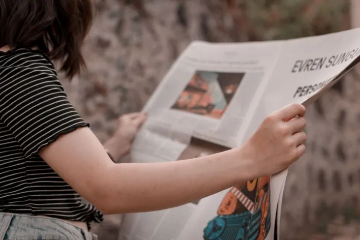 A woman holding a newspaper while sitting outside, enjoying a leisurely read.