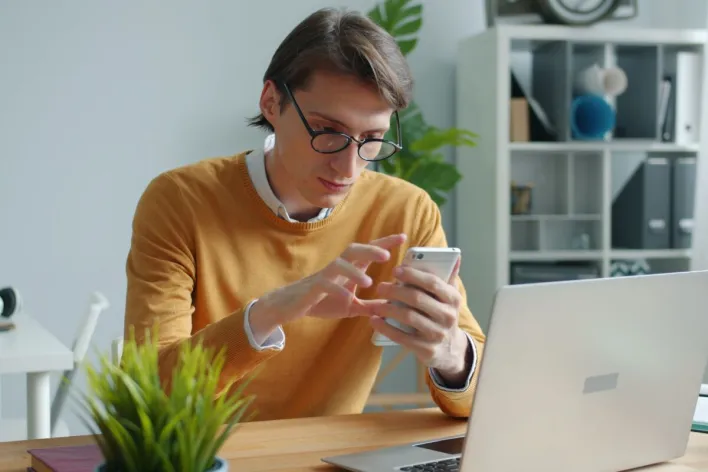 A young adult working on a smartphone and laptop in a modern office.
