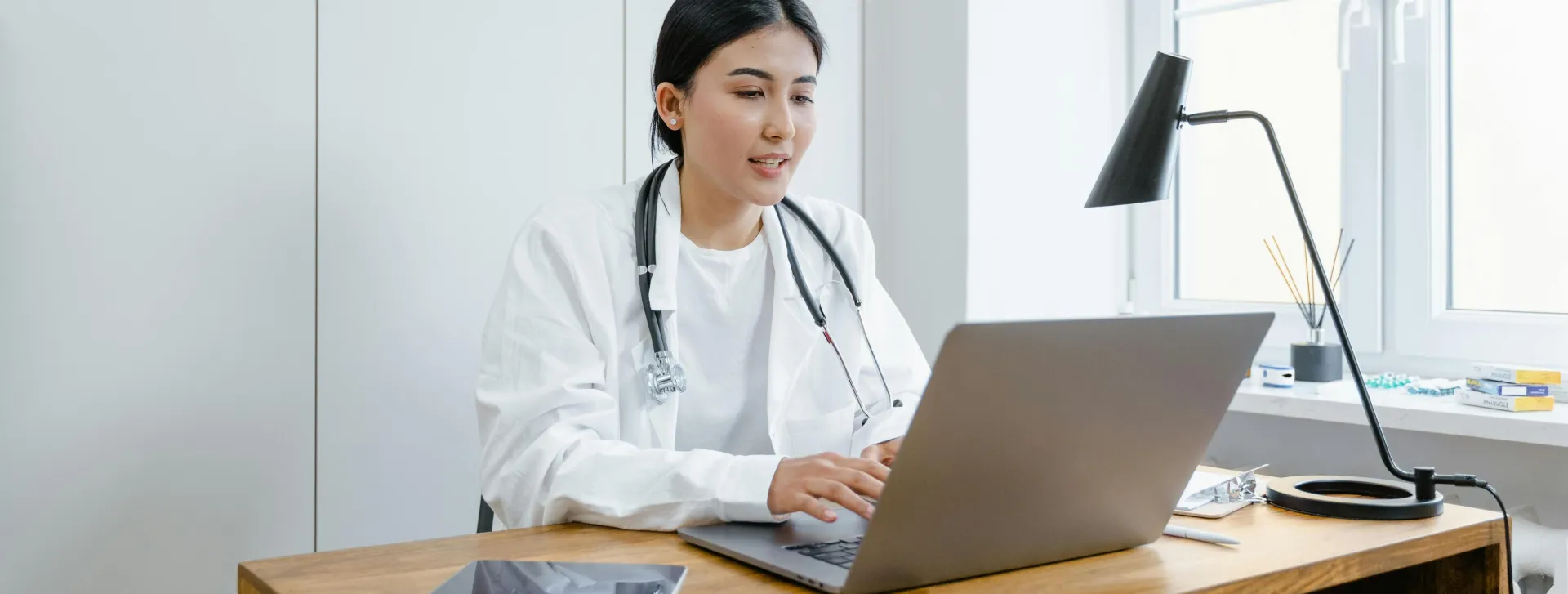 A female doctor in a white coat uses a laptop for an online consultation from her office.