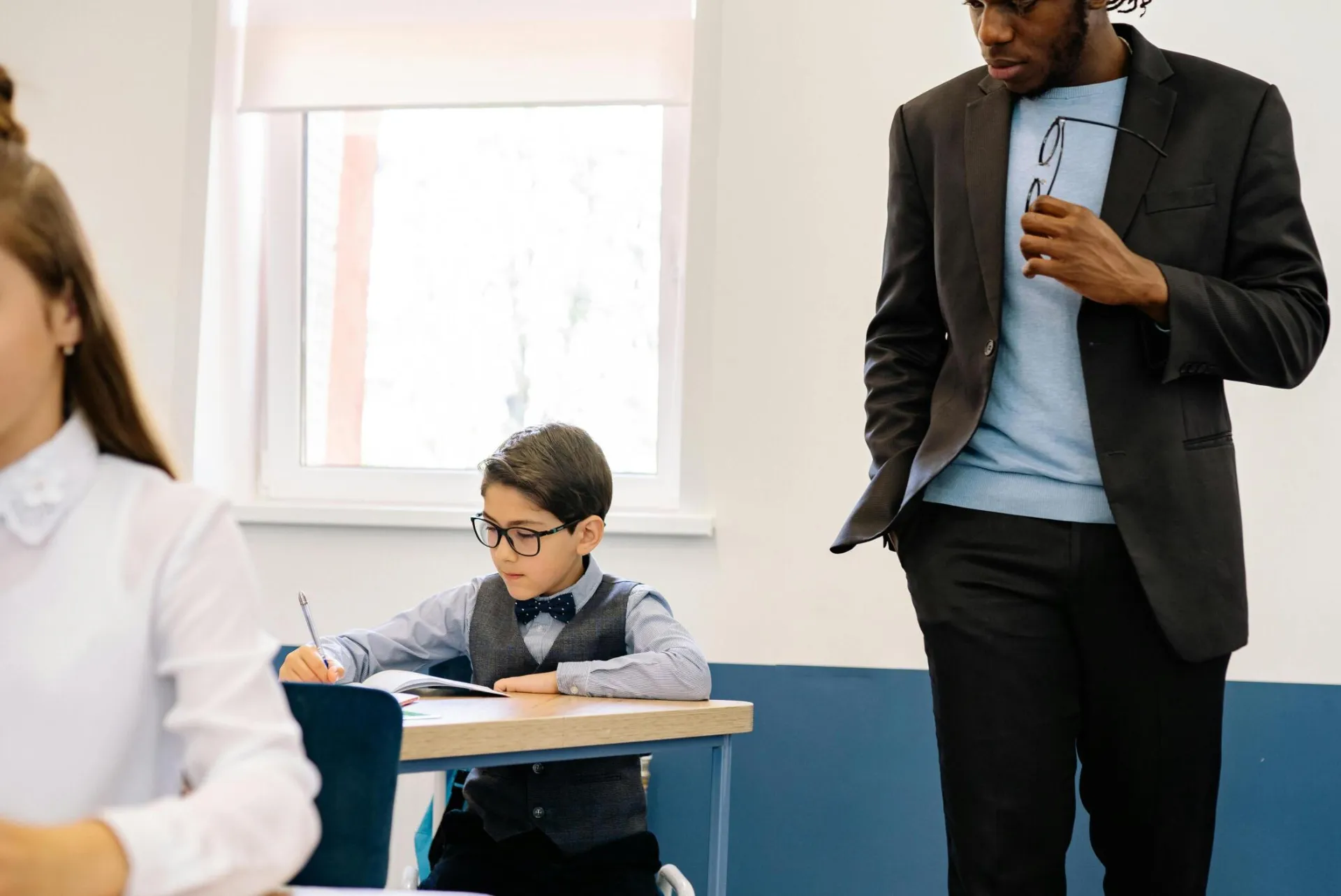 A teacher in a black suit supervises young students working at desks in a classroom.