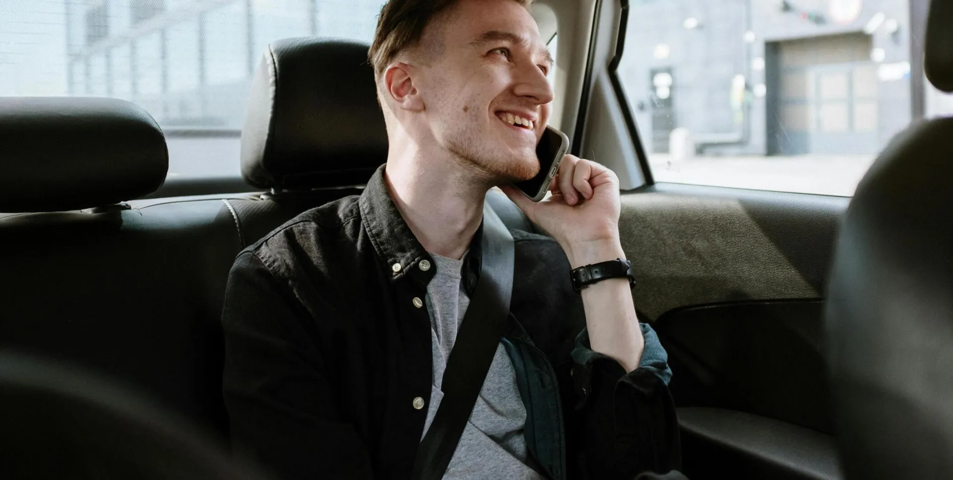 Smiling young man enjoying a phone call while seated in the back seat of a car during a daytime ride.
