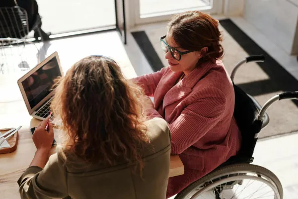 Two women working on laptops, one using a wheelchair, collaborating in a modern café.