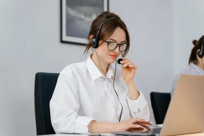 Young woman in glasses and headset providing customer support at a laptop in an office setting.
