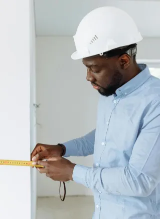 Construction worker measuring a wall indoors, wearing a white hard hat and blue shirt.