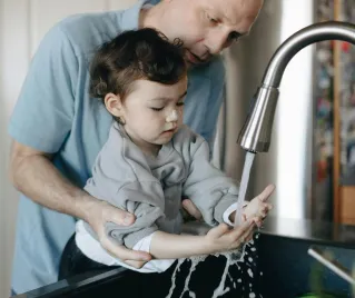 A father tenderly helps his child wash hands in the home kitchen sink.
