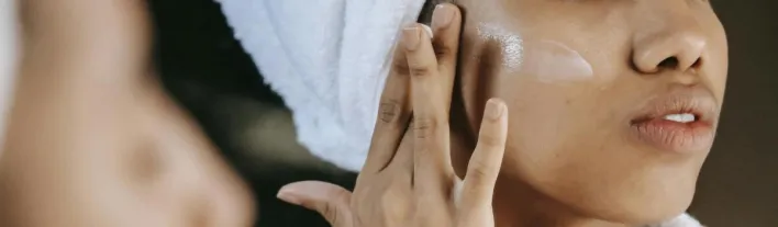 Crop unrecognizable young ethnic female in terry robe applying moisturizing cream on cheek while reflecting in mirror