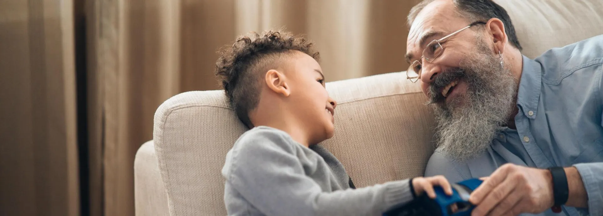 A joyful grandfather and grandson sharing a playful moment on the couch, indoors.