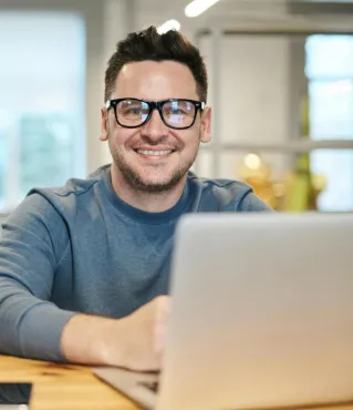 Happy man wearing glasses working remotely on laptop in modern office environment.