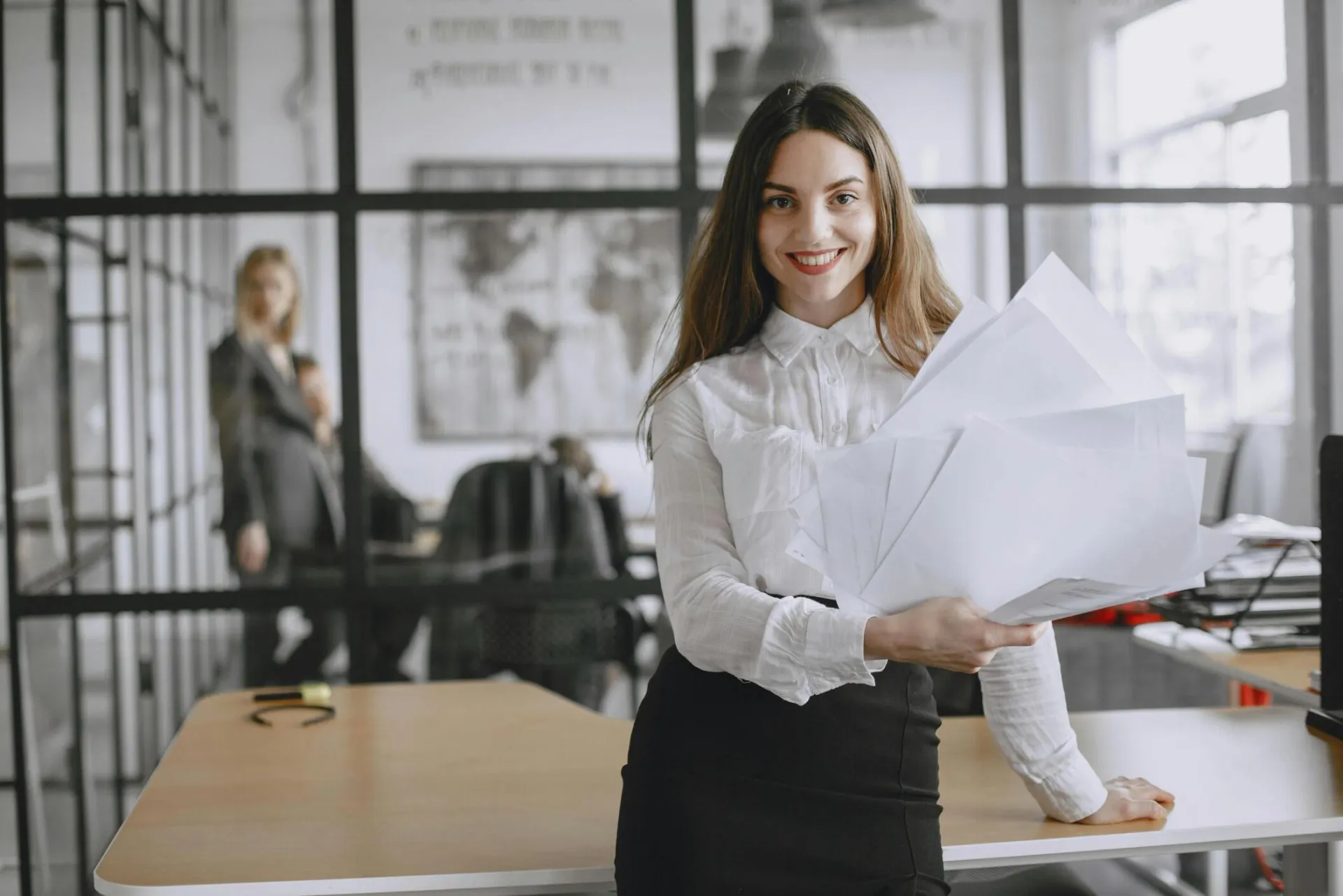 Confident young woman in white shirt smiling while holding papers in a modern office environment.