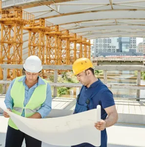 Two engineers on a construction site examining blueprints with steel structures in the background.