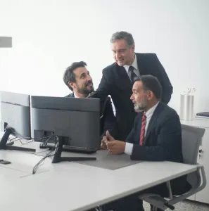 Three business executives in suits collaborating around dual monitors in a modern office setting.