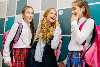 Three girls in school uniforms laughing and talking by lockers, indoors.