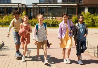 A diverse group of children walking outdoors, carrying backpacks and a basketball, heading to school on a sunny day.