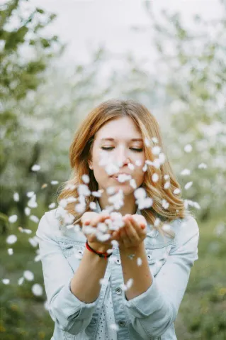 A young woman with wavy hair blows cherry blossom petals in a scenic orchard during springtime.