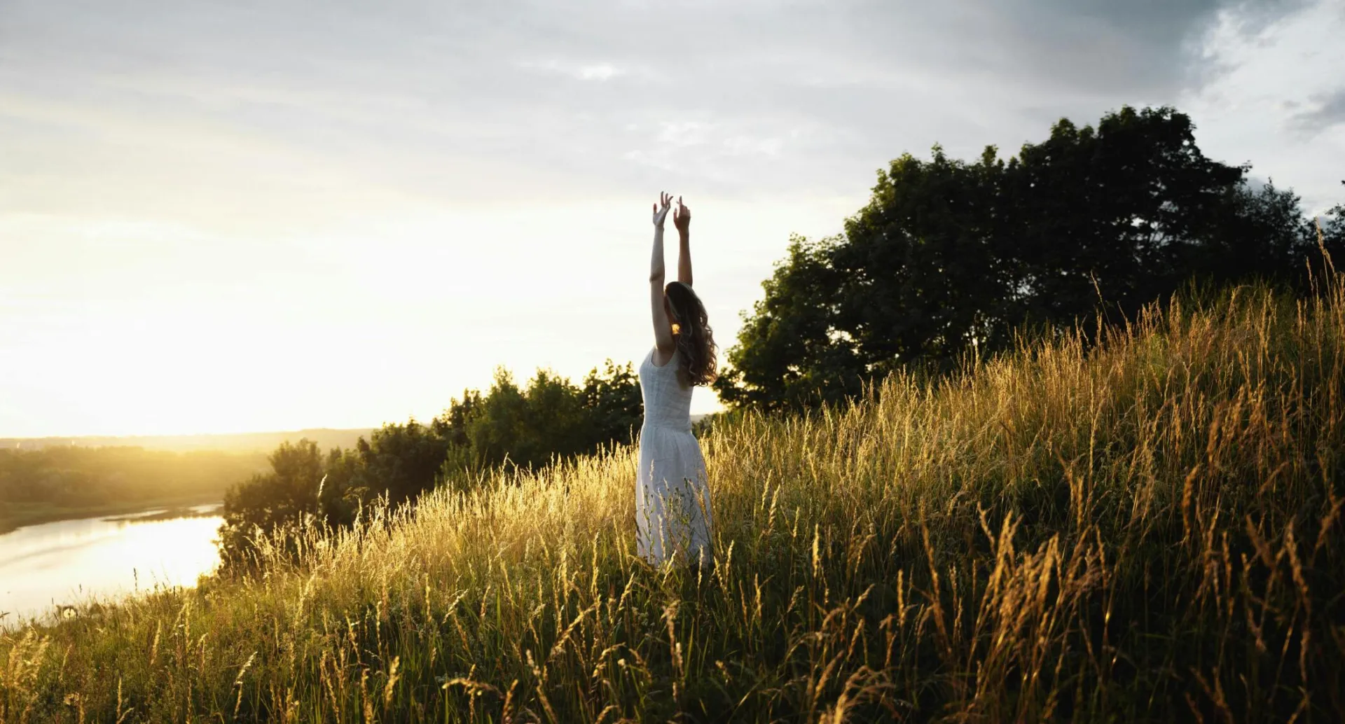 A woman in a white dress enjoys the sunrise in a scenic grassy field by the water.