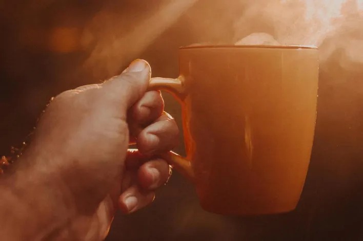 A warm hand holding a steaming cup of coffee against a sunny backdrop.