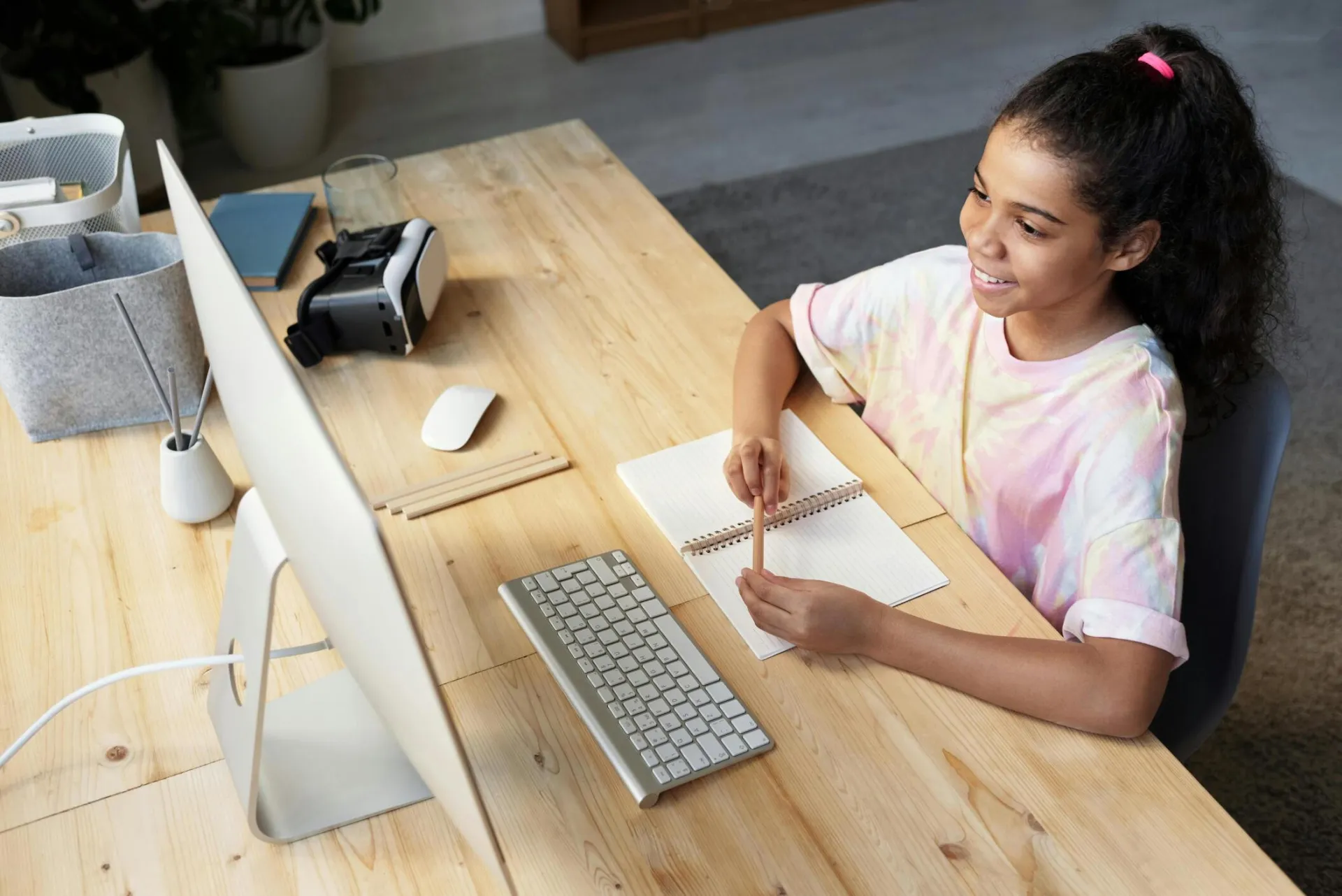 A young girl smiling while studying online at home, using a computer and taking notes.