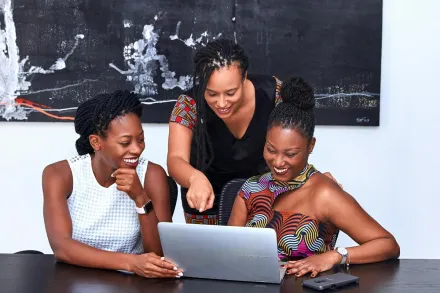 Three African American women collaborating at a laptop, focused and happy during a meeting indoors.
