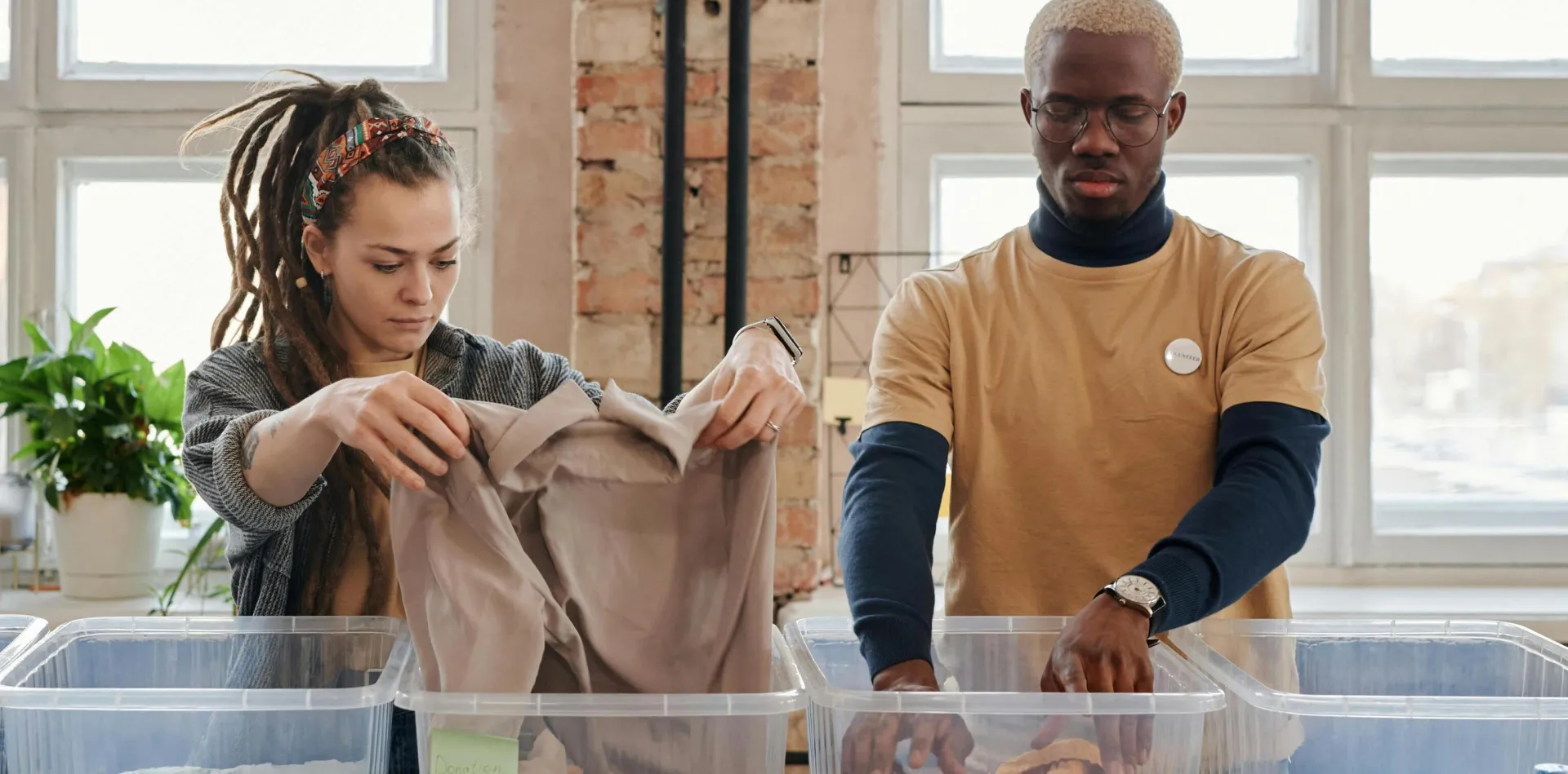 Two volunteers organize clothing and food donations indoors during daylight.