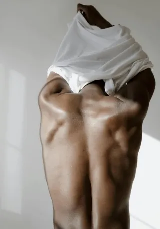 Rear view of a muscular man lifting shirt, showcasing toned back muscles, in a bright setting.