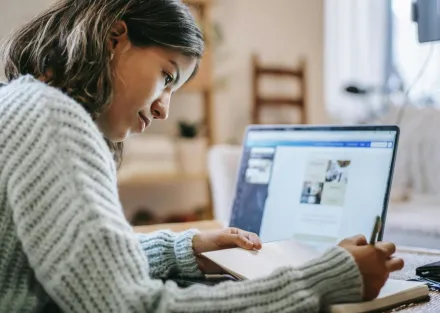 Side view of concentrated young Latin American female freelancer in casual clothes using laptop while taking notes in notebook with pen at table near ring light with phone in room