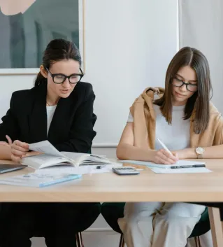 Two women working together with books and a laptop, focused on learning.