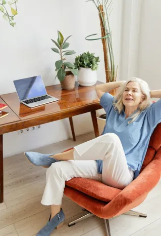 Senior woman in modern office setting, relaxing in a chair with a laptop on a desk.