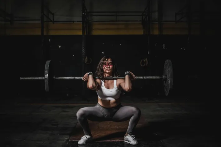 A strong female weightlifter squats with a barbell in a dimly lit gym, showcasing power and fitness.