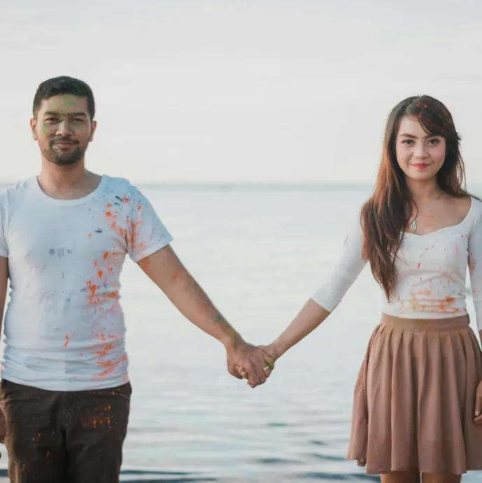 A cheerful couple holding hands by the sea, enjoying a playful day with paint splashes.
