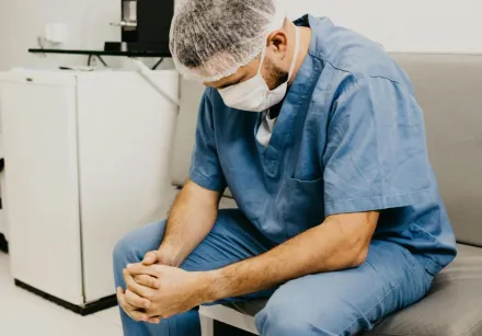 A male doctor in hospital attire sitting pensively, representing healthcare challenges.