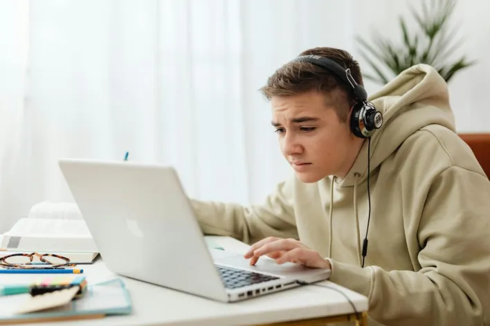Teen wearing headphones studies online with a laptop in a cozy indoor setting.