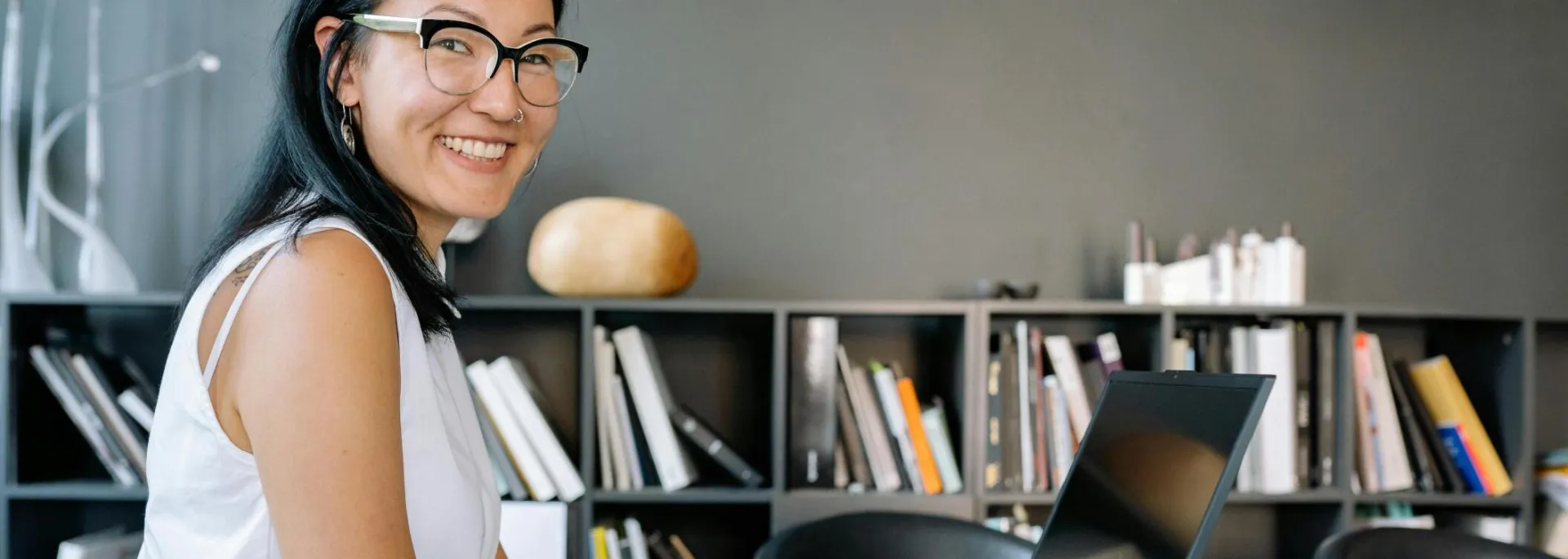 Cheerful businesswoman using laptop at a desk in a stylish office setup with bookshelves.