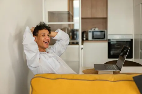 Casually dressed man enjoying leisure time at home with an open laptop, expressing happiness and relaxation.