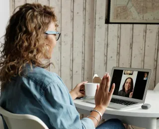 A woman in a blue shirt interacts on a video call using a laptop in a cozy home setting.