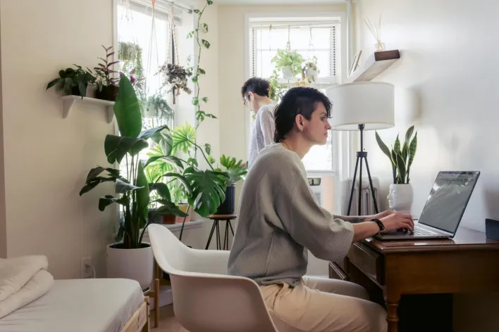 Attentive female surfing internet on netbook while sitting in armchair against potted plants and partner in house