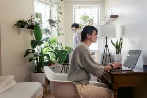 Attentive female surfing internet on netbook while sitting in armchair against potted plants and partner in house