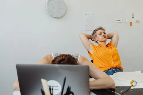 Two women in office, one stressed with head on desk, another relaxed. Modern workplace dynamics.
