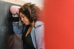 Side view of tired young black female with dark curly hair in sportswear and protective gloves leaning on wall with closed eyes after hard training in gym