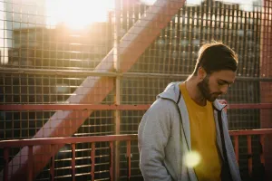 Young upset unshaven male in casual apparel looking down near grid fence in back lit in town