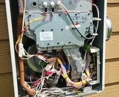 Tankless water heater on outside of home mounted on a wall. Front cover is off exposing the inside wires & components.