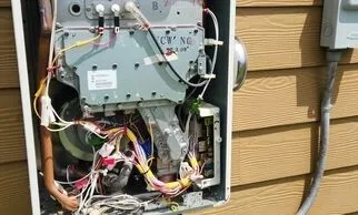 Tankless water heater on outside of home mounted on a wall. Front cover is off exposing the inside wires & components.