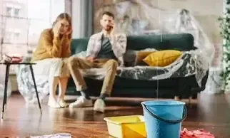 Man and women sitting on couch with looks of frustration as leak in ceiling fills plastic bucket.