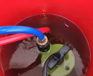 Red 5 gallon bucket with bump pump in dirty flush liquid. Red and blue hose end into bucket and other end out.