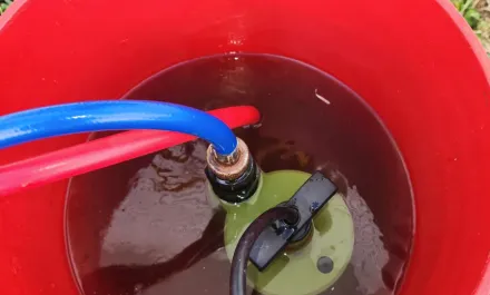 Red 5 gallon bucket with bump pump in dirty flush liquid. Red and blue hose end into bucket and other end out.